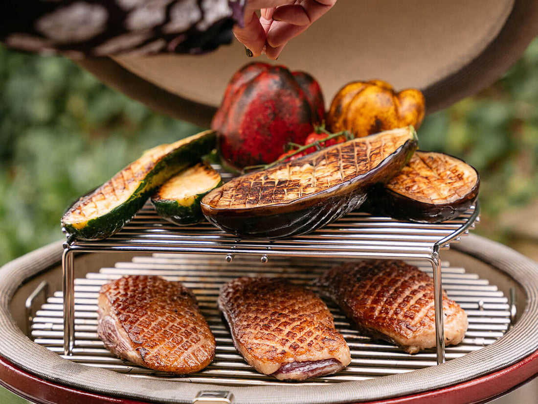 Cuisson en deux niveaux dans un kamado Kokko avec magrets de canard saisis sur la grille inférieure et légumes grillés sur la grille surélevée, illustrant la polyvalence de cuisson.