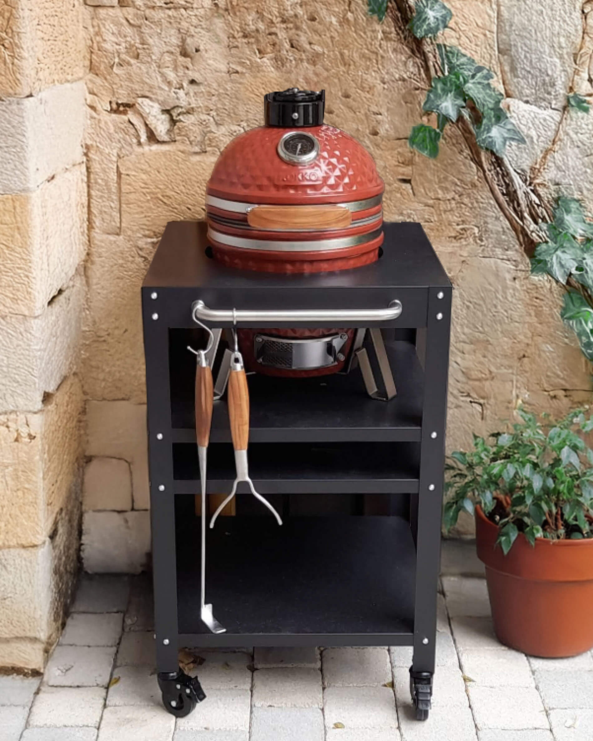 Kamado Kokko rosso in ceramica posizionato su un carrello nero con ruote, dotato di ripiano superiore, vano portaoggetti aperto e utensili appesi sul davanti, presentato in un cortile in pietra.