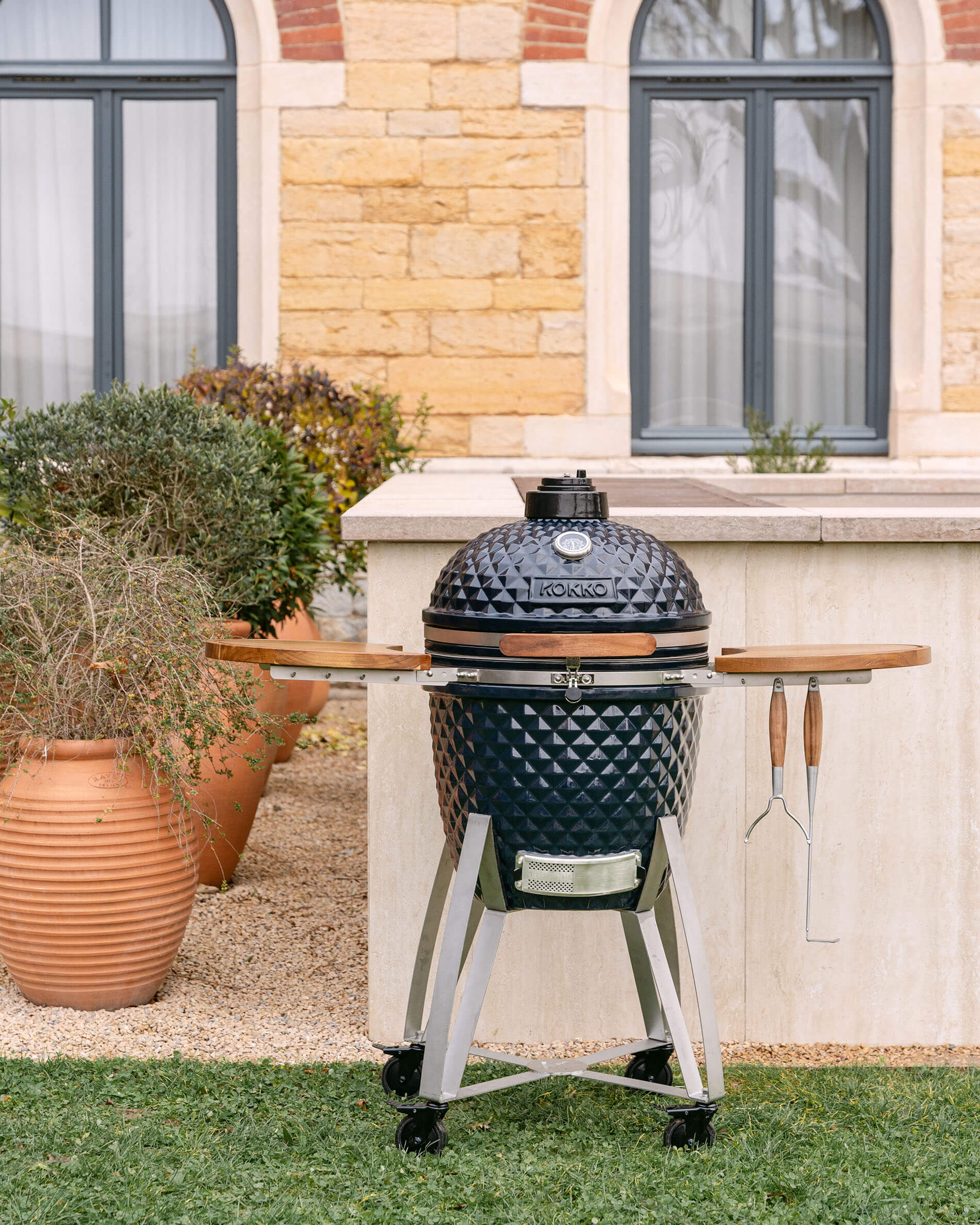 Gesamtansicht des Kokko Kamado auf mobilem Wagen mit klappbaren Ablagen und hängenden Grillutensilien, im Outdoor-Einsatz.