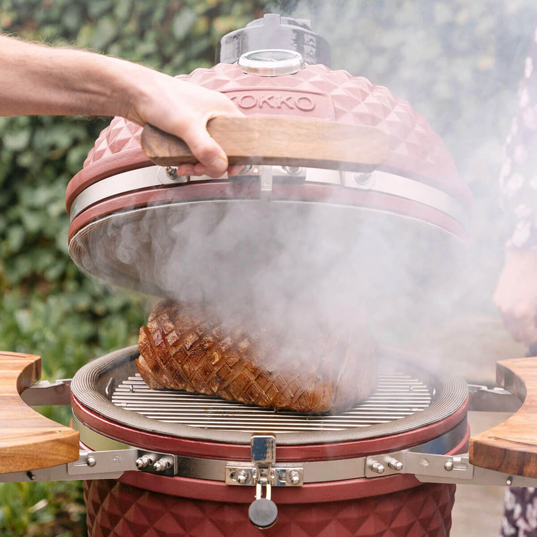 Une main soulève le couvercle d’un kamado Kokko rouge oxyde, laissant apparaître une pièce de viande grillée posée sur la grille en inox. Une importante fumée s’échappe de la cuve, mettant en valeur la cuisson au feu de bois, le joint périphérique et les plateaux latéraux en bois.