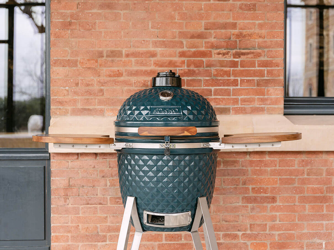 Kamado Kokko de couleur vert océan, installé en extérieur devant un mur en briques. Le barbecue est fermé, avec un couvercle en céramique texturée, un thermomètre intégré sur le dessus, une poignée en bois, un loquet de sécurité en inox et deux plateaux latéraux en bois d’acacia. L’ensemble repose sur un support métallique avec bac de ventilation inférieur visible.