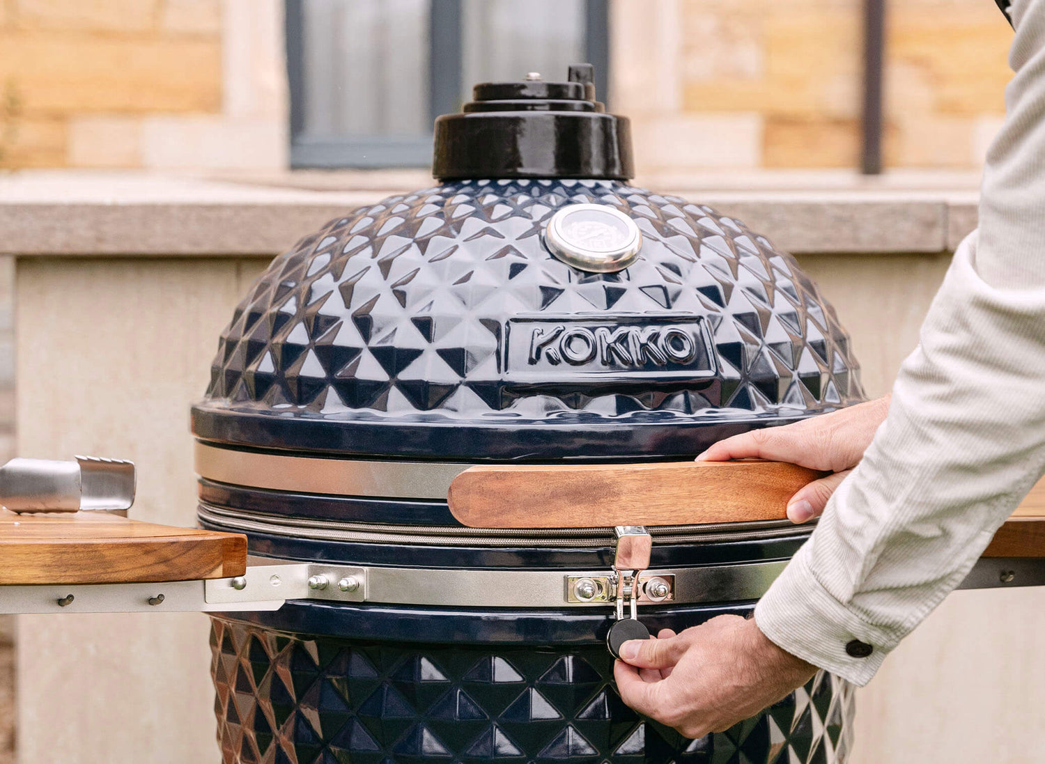 Main actionnant le loquet de sécurité d’un kamado Kokko noir, avec couvercle en céramique texturée, poignée en bois et thermomètre intégré, en situation d’utilisation en extérieur.