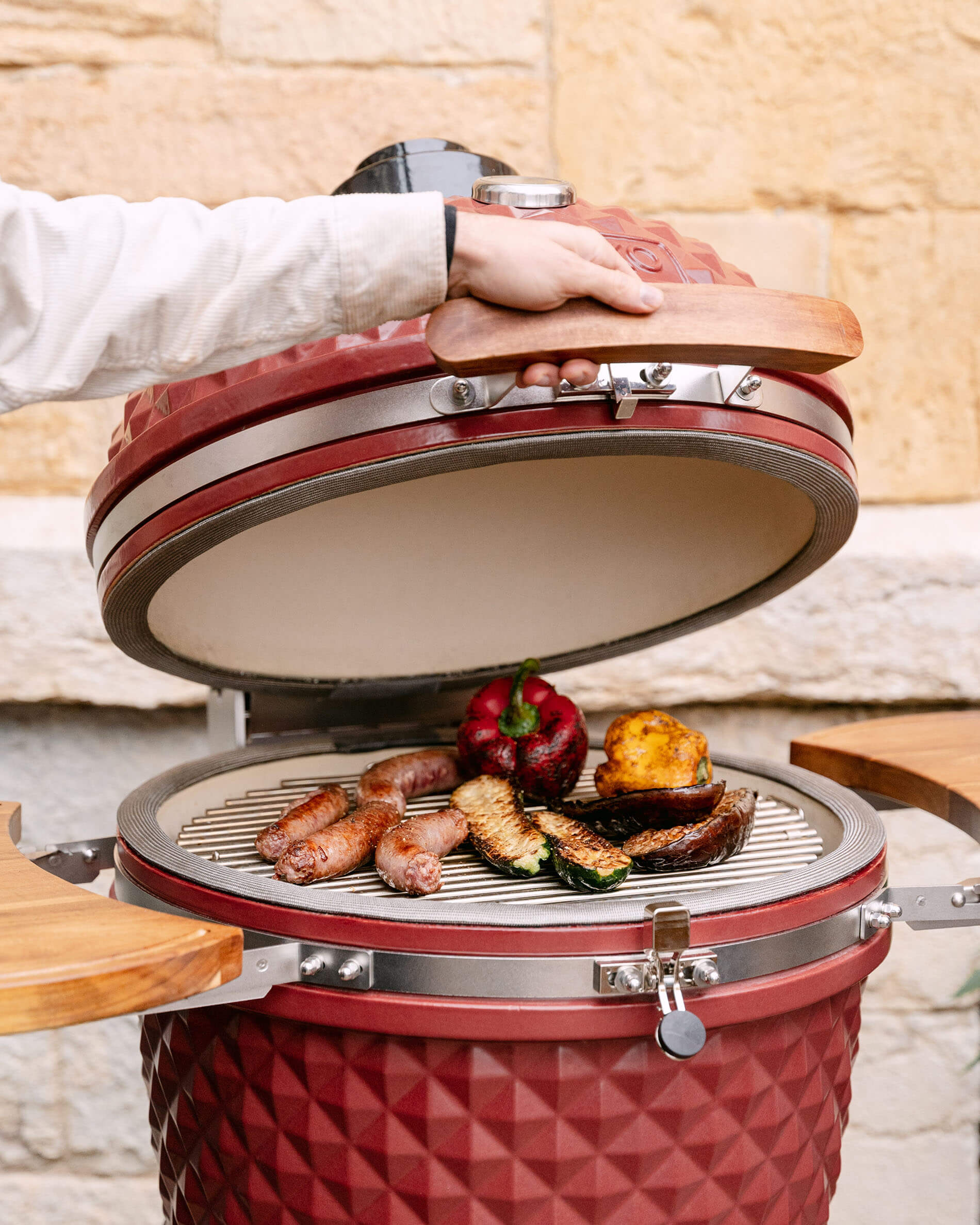 Kamado Kokko 22 pollici in ceramica rosso ossido aperto, con salsicce e verdure in cottura sulla griglia.