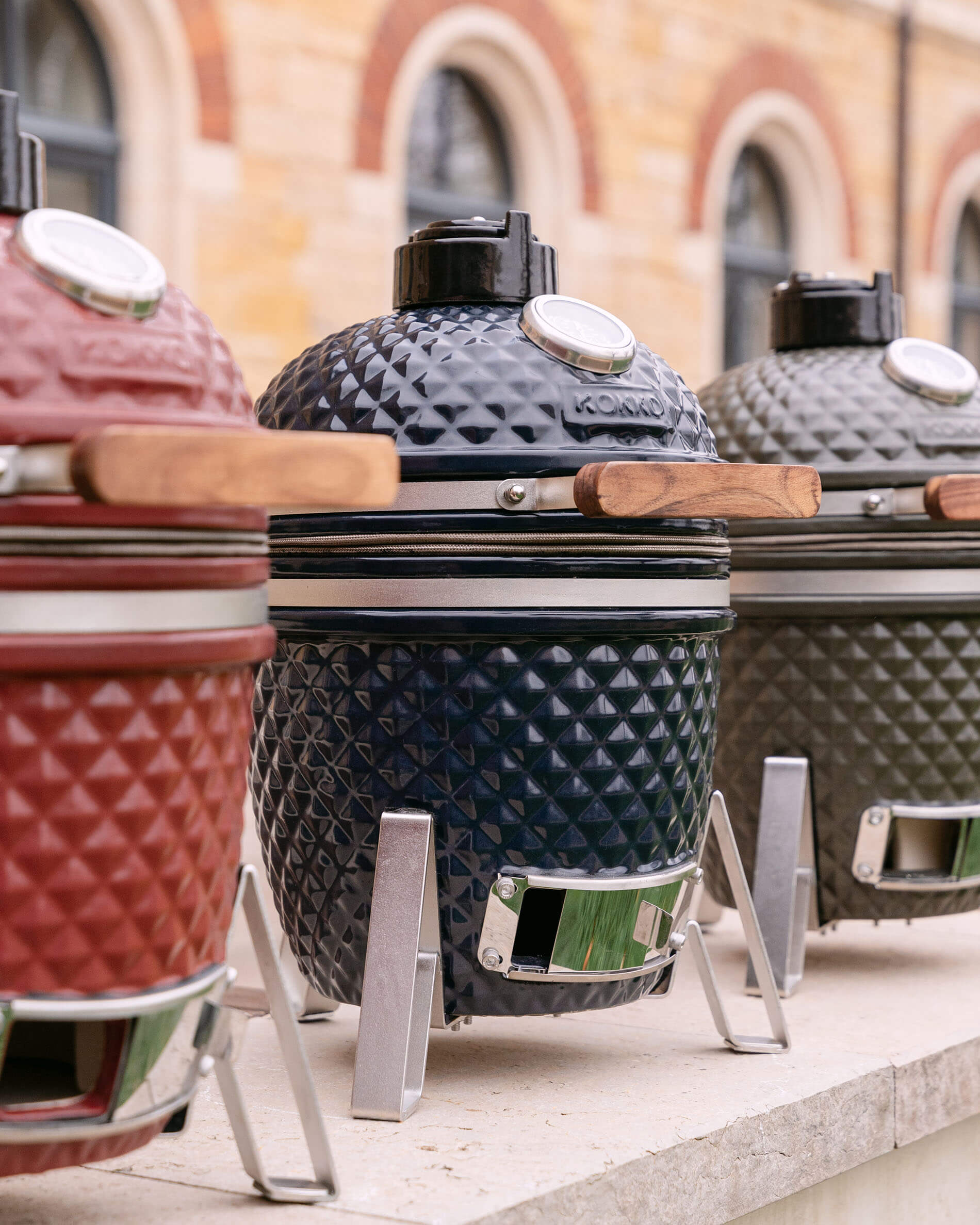 Reihe von Kokko Kamado-Grills aus strukturierter Keramik, in verschiedenen Farben mit Holzgriffen und Metallgestellen.