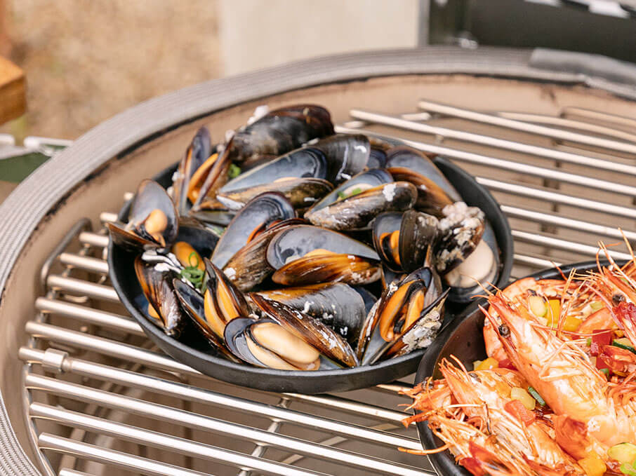 Moules cuites dans un plat posé sur la grille d’un kamado Kokko, accompagnées de crevettes, illustrant une cuisson indirecte de fruits de mer en extérieur.