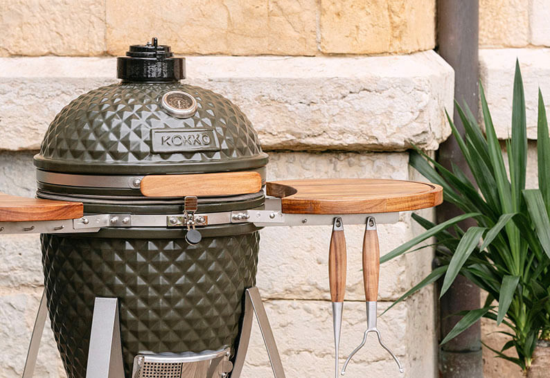 Kamado Kokko nouvelle édition installé en extérieur, avec céramique noire texturée, tablettes en acacia et châssis métallique.